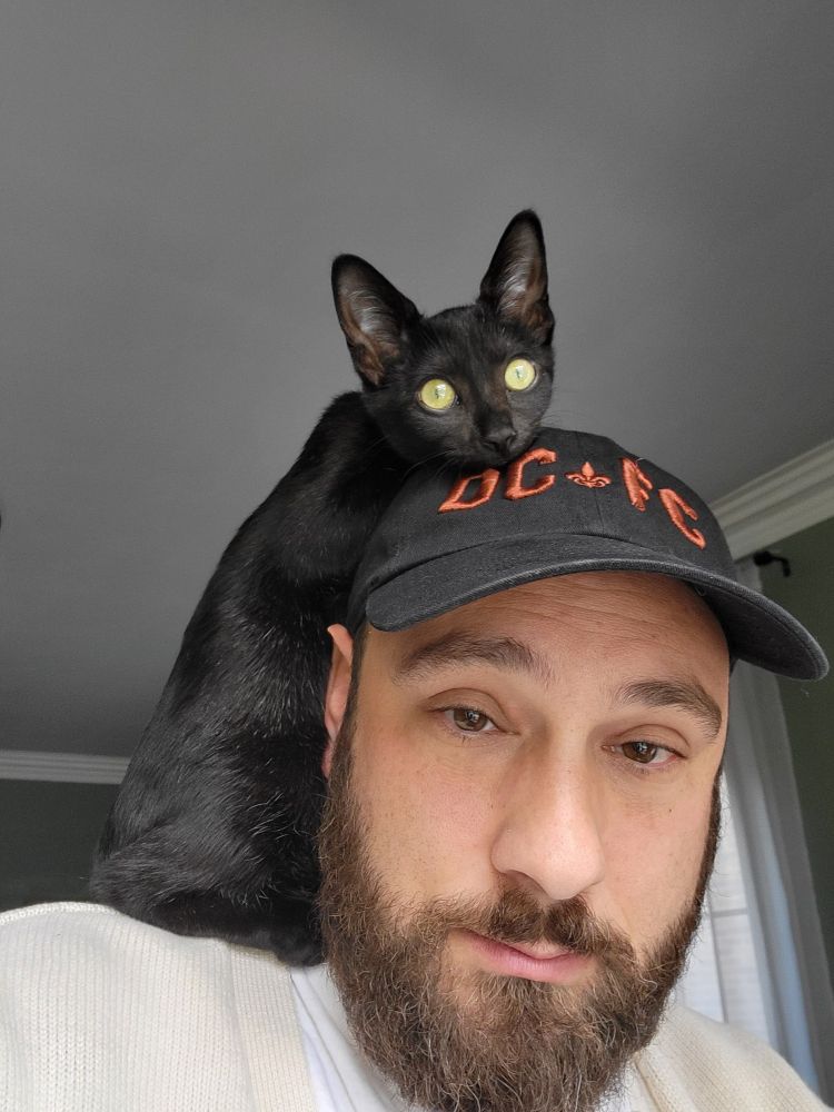 A picture of a man (myself) with a black cat on his shoulders. The cat is laying her head on his hat.