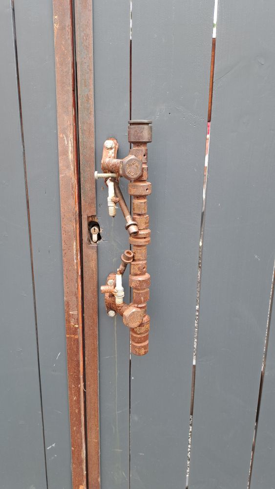 Ein Tor aus grauen Latten mit metallischen, rostfarbenen Kanten. Darauf ein großer vertikaler Griff aus unterschiedlichen Schrauben und Verbinderstücken, ebenfalls rostfarbenen. 