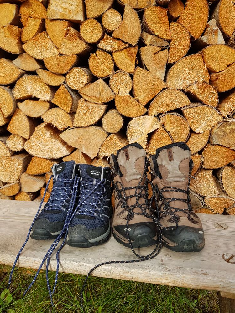 Ein Paar blaue und ein Paar braune Wanderschuhe, die auf einer Holzbank vor einem Stapel frischer Holzscheite stehen. 