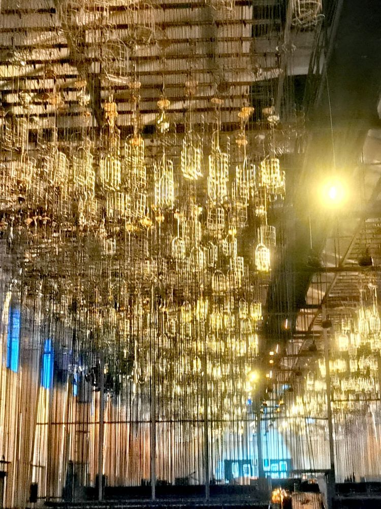 Viele Körbe aus Metall, die an Ketten von der Decke hängen, werden von hellem Licht angestrahlt. An den Seiten hohe Wände, die oben in Abschnitten blau beleuchtet sind. 