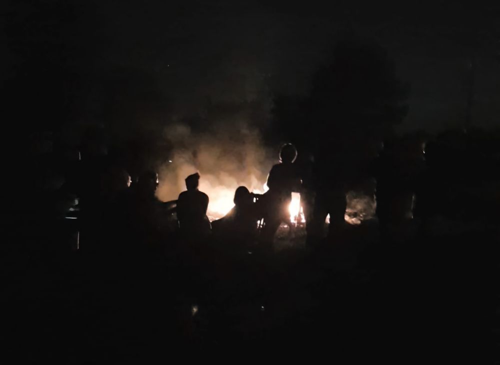 Eine Gruppe von Menschen steht oder sitzt mit dem Rücken zum Betrachter um ein Feuer 