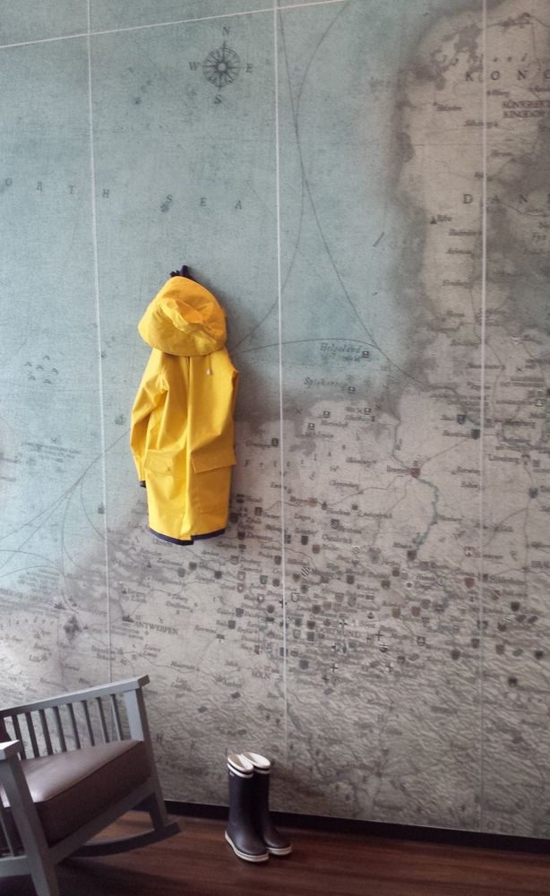 Eine Wand, bedruckt mit einer historischen Landkarte der deutschen Nordseeinseln in Sepia und blassblau. An der Wand hängt ein gelber Regenmantel (Friesennerz), davor steht ein Paar blaue Gummistiefel auf braunem Holzboden. Links unten im Bild ein Teil eines grau-braunen Schaukelstuhls.