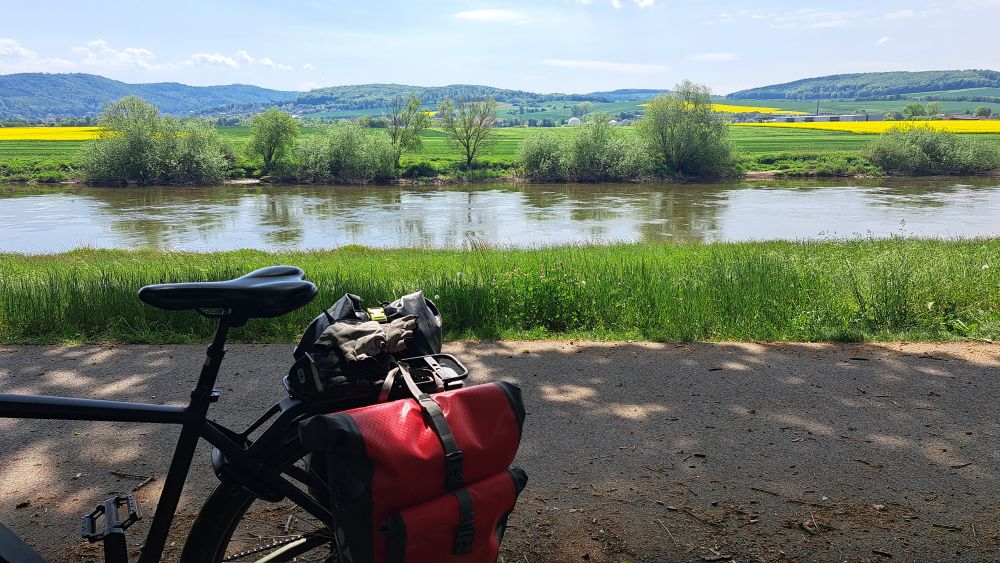 Die hintere Hälfte eines schwarzen Herrenrads steht an einem Weg. Am Gepäckträger rote Fahrradtaschen. Hinter dem Weg verläuft ein Grünstreifen, der bis an die Weser reicht. Am gegenüberliegenden Ufer einige Sträucher, dahinter grüne Wiesen und gelbe Rapsfelder. Im Hintergrund dunkelgrüne Hügel. 
