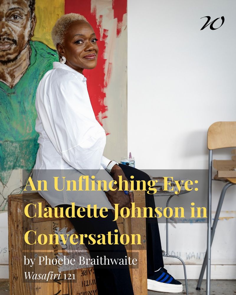 Photo of a Black woman in a white shirt and black trousers. She has cropped blonde hair and sits on a wooden crate. In the background is a painting of a Black man in a green top.
