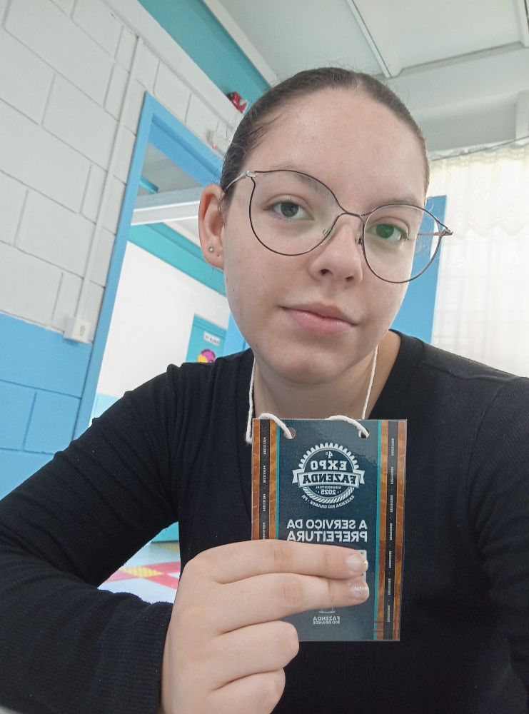 Eu (blusa preta, cabelo castanho amarrado, óculos redondo) com crachá da prefeitura, fundo parede branca e porta azul claro 