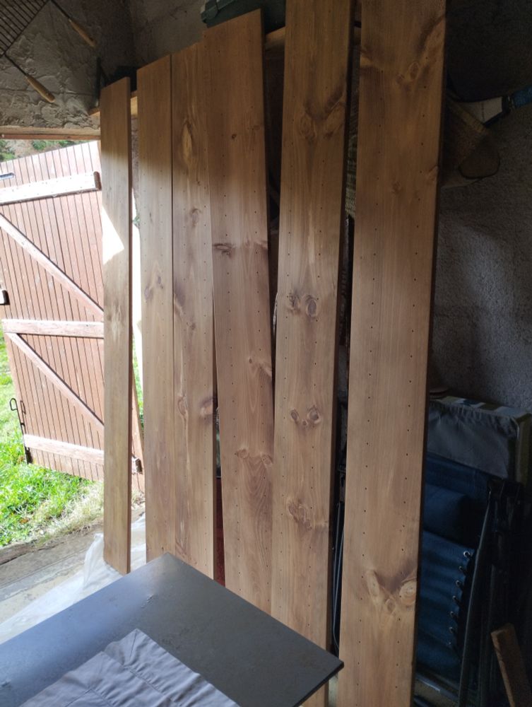 Montants de bibliothèques (6 en tout) en pin, teintés et huilés, posés à la verticale pour sécher dans un garage dont la grande porte est ouverte, laissant deviner un peu du bazar à la lumière du jour