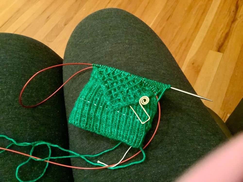A woman’s lap,gray sweatpants over hardwood floor. In the forgeound are circular knitting needles with a red cable and green yarn leading to an intricately cabled and twisted sock cuff.