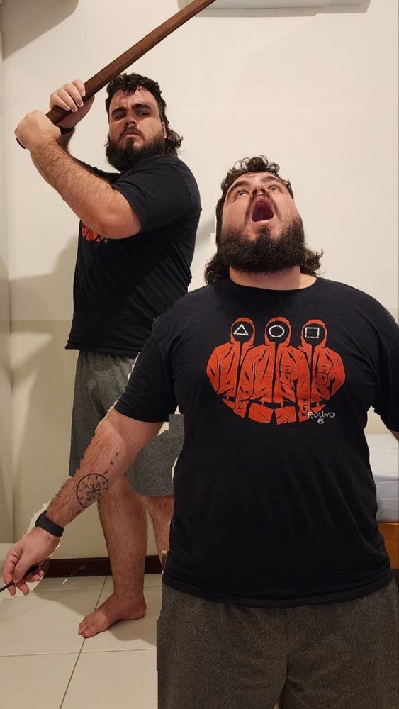 A imagem é uma imagem de referência, mostrando eu como duas pessoas diferentes. O da esquerda está em pé, com expressão séria e um bastão de madeira levantado em posição de ataque. O da direita está à frente, com a cabeça inclinada para trás e a boca aberta, como se estivesse gritando ou reagindo, segurando um objeto pequeno na mão. Ambos usam camiseta preta com estampa de três figuras encapuzadas vermelhas com símbolos geométricos e a palavra "ROUND 6", e shorts cinza.