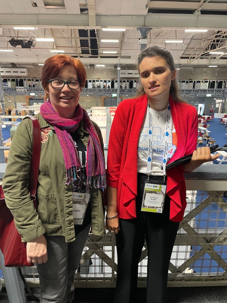 Elizabeth, a white woman with long brown hair, poses with Catherine Lyons, a white woman with short red hair, on a balcony overlooking a large conference room. Catherine, on the left, wears glasses, an olive jacket, pink scarf, red purse, and grey jeans. Elizabeth, on the right, wears a white Communication 4 ALL t-shirt, red jacket, and black pants, and holds her iPad. Both wear conference nametag lanyards.
