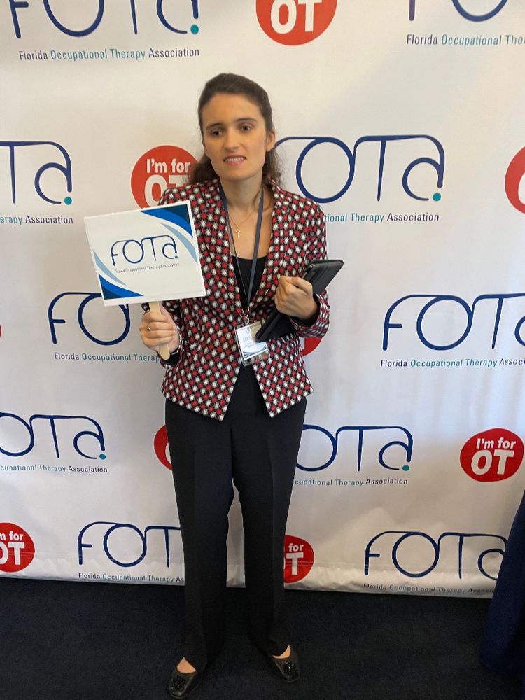 Elizabeth, a white woman with long brown hair half-up half-down, poses in front of a white backdrop with repeating FOTA (Florida Occupational Therapy Association) logos and red circles with white text that reads “I’m for OT!” Elizabeth wears a navy, white, and red checker-patterned blazer with a black blouse, slacks, and flats, with a conference nametag lanyard. She holds a sign with the FOTA logo on it in one hand, and her iPad in the other.