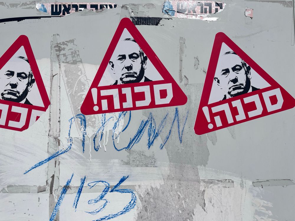 Drei rote dreieckige Aufkleber mit einem Porträt von Benjamin Netanjahu und dem hebräischen Wort „סכנה!“ (Gefahr!) sind auf eine Wand oder eine öffentliche Fläche geklebt. Die Wand ist mit weiteren Graffiti und Aufklebern bedeckt.