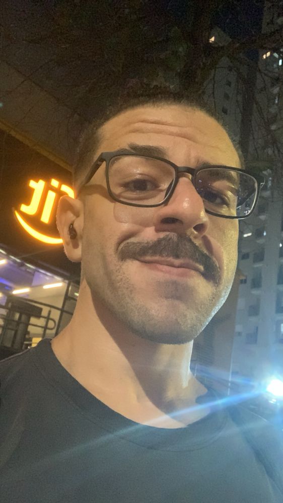 Selfie tirada de baixo pra cima, homem branco de óculos olhando pra câmera, e com um sorriso de canto de boca