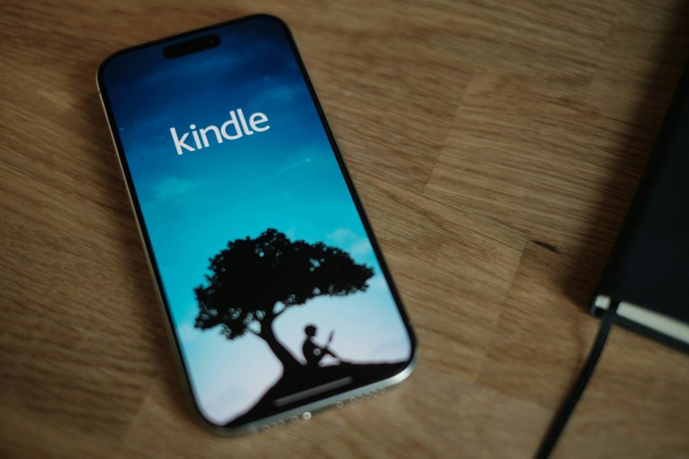 Imagem de um smartphone moderno apoiado sobre uma superfície de madeira clara. Na tela do aparelho, está aberto o aplicativo Kindle, exibindo sua tela inicial característica: um fundo em tons de azul e ciano que simula um céu ao entardecer, com a silhueta preta de uma grande árvore e, embaixo dela, a figura de uma criança sentada lendo um livro. No centro superior da tela aparece a palavra "kindle" em letras brancas. À direita da imagem, parcialmente visível, há um caderno preto com elástico.