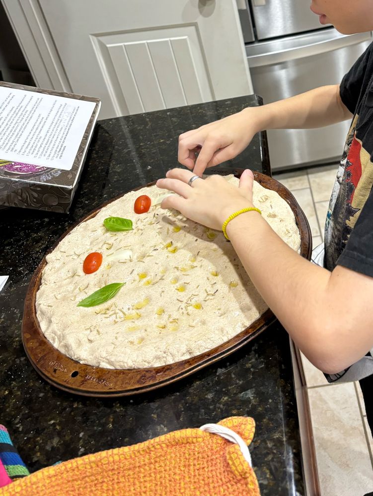 B decorating the focaccia with cherry tomatoes and onion pieces to look like mushrooms and fresh basil leaves. 