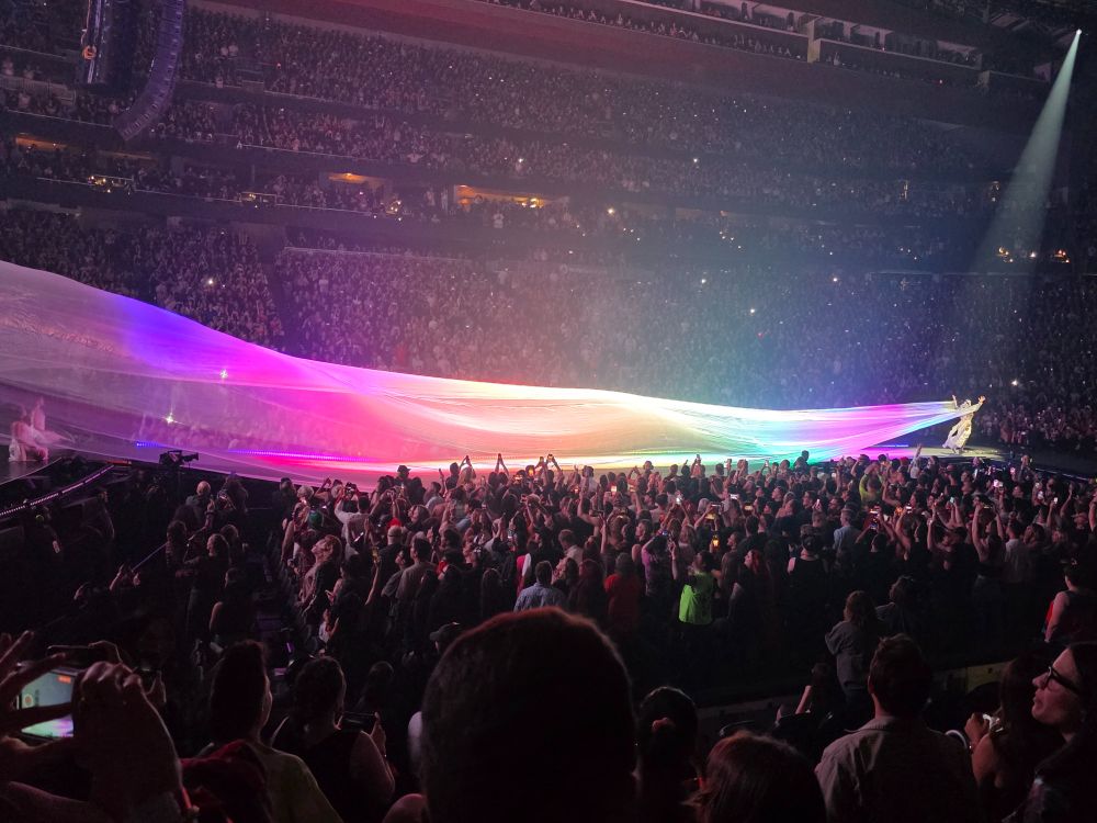 A long-view scene of a concert in an indoor stadium with the lights down, the seats filled with people, with Lady Gaga wearing an extremely long, flowing veil bathed in rainbow lights.