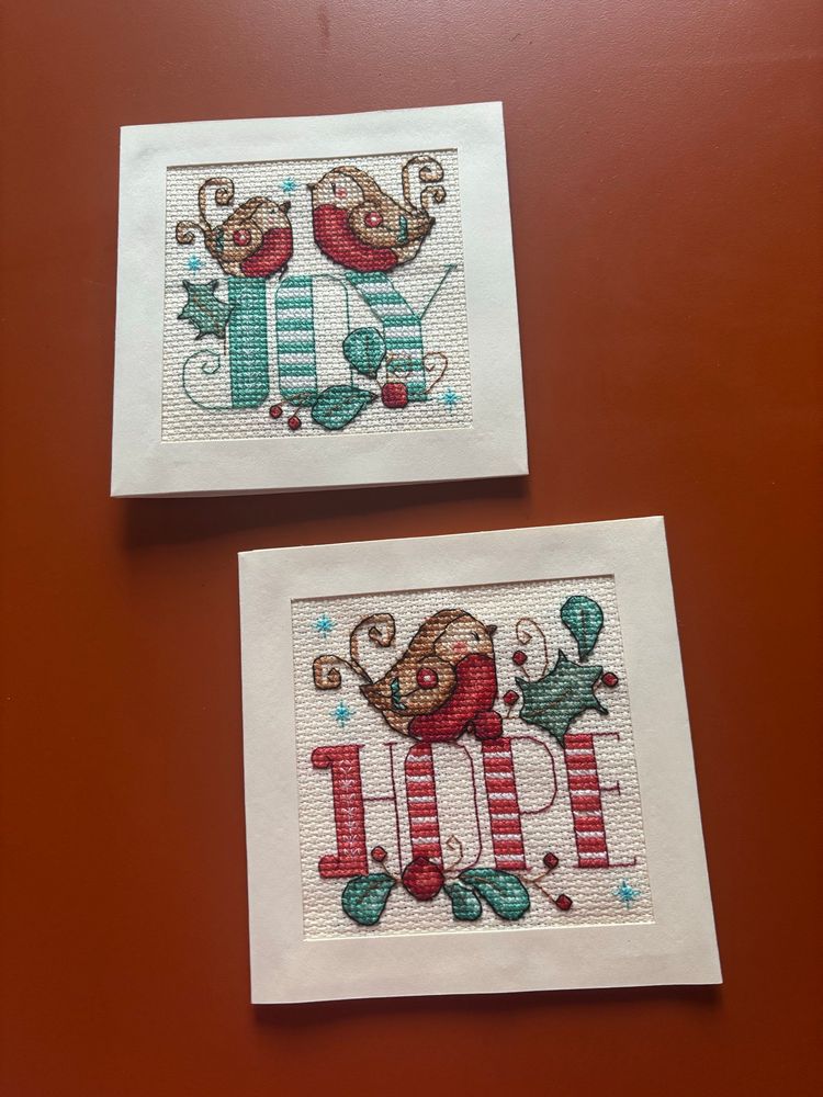 Two cross stitched Christmas cards on an orange tabletop. Both cards have a robin design and one says hope and the other says joy. 