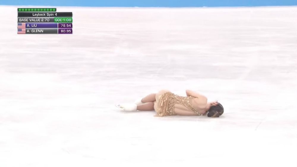 a figure skater (alysa liu) in a gold costume lying resignedly on her side on the ice, facing away from the camera