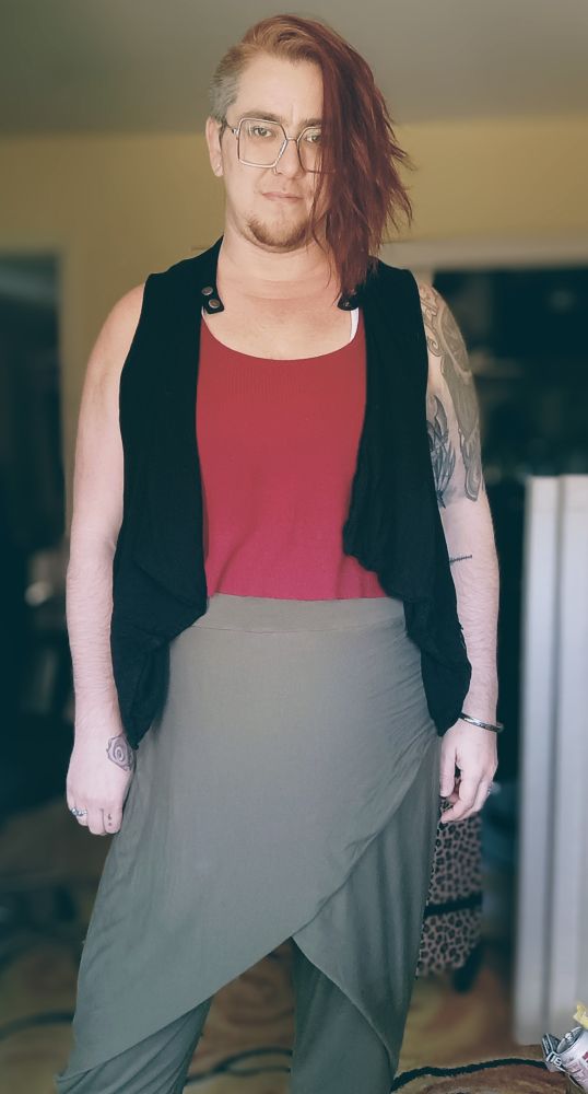 Marin stands in a neutral pose facing the camera, wearing a black vest, red tank, and green wrap pants. Square glasses rest behind long red hair and above a goatee.