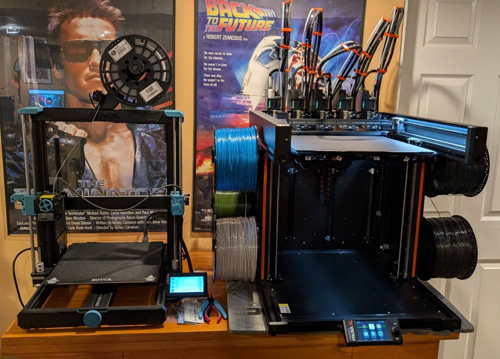 Photo of two 3D printers side by side, one from Sovol on the left, and a new Prusa XL holding five different filaments on the right. In the background on the wall are a Terminator and Back to the Future posters.