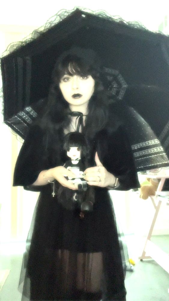A half-body photo of myself standing and holding the aforementioned doll (described in the previous post). I am pale with long black hair and swept bangs. I have dark lipstick and eye makeup, looking towards the camera with a neutral expression. I am wearing a black faux velvet & cropped cape, secured by a black ribbon at my neck. I hold Nikola, my doll, at chest-height. She is in a sitting position and continues to stare off to the side all creepy like. My outfit is completed with a layered skirt, sheen and of a black mesh fabric. I also carry a parasol, adorned with white and black lace designs.
