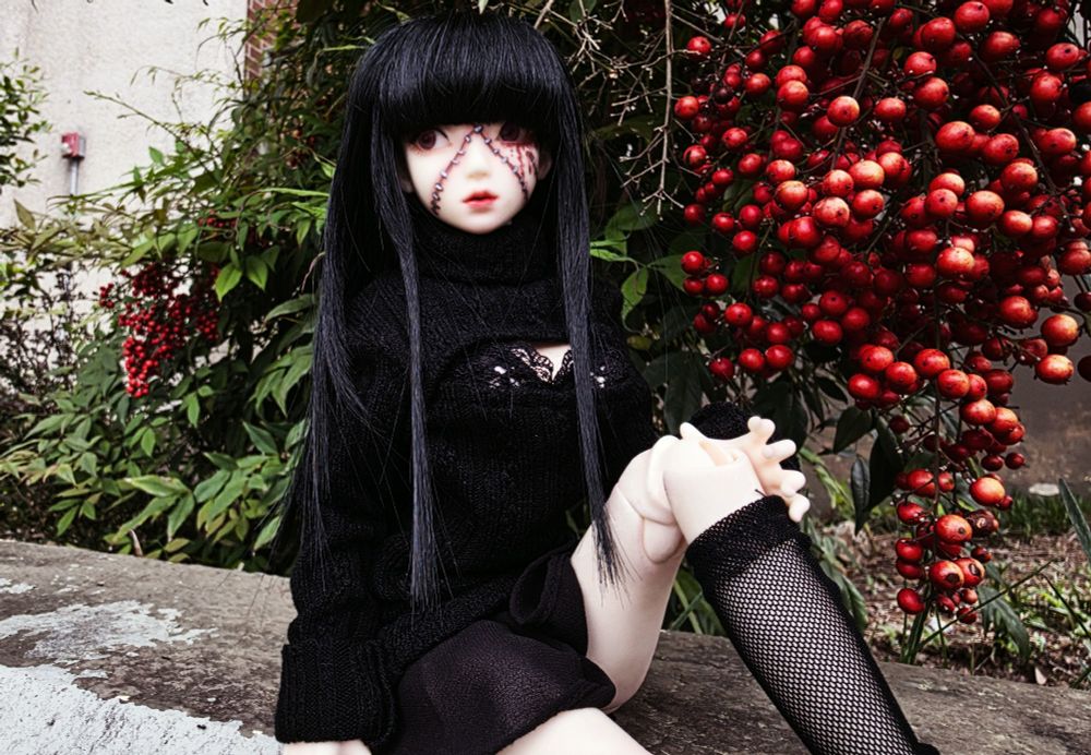 A digital photo of Hiromi sitting on the same concrete ledge. Now the angle showcases a bustle of red berries behind her. She is sitting with one knee propped up, her hand resting on the side of her leg.