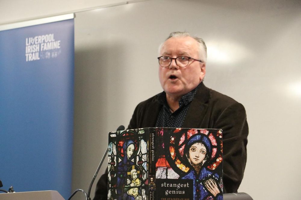 Gerry Molumby speaking at a lectern which has a book on stained glass propped up on it 