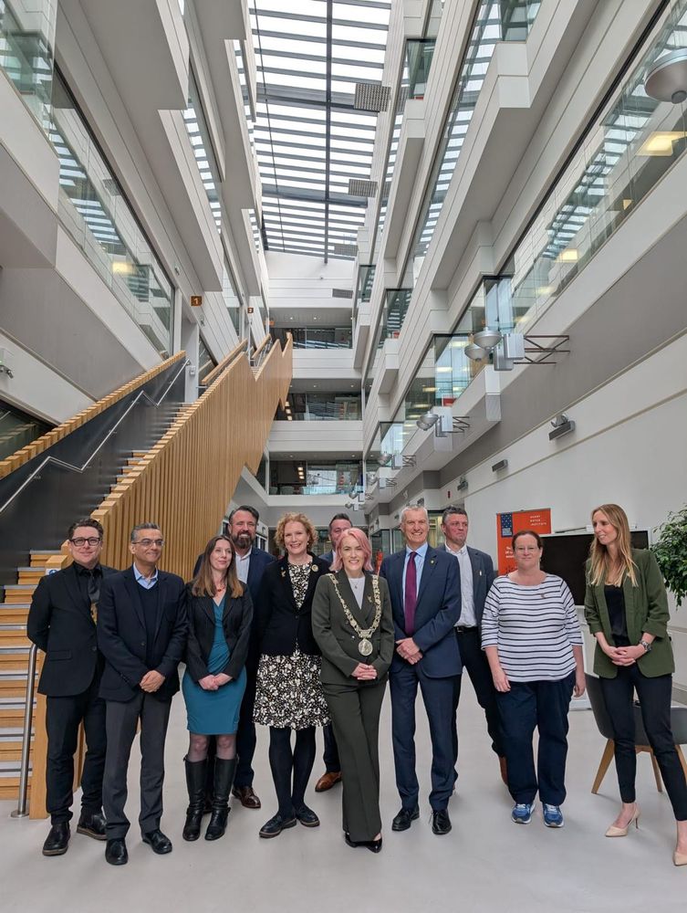 Lord Mayor of Dublin, Emma Blain, and University of Liverpool VC Tim Jones, standing in the centre of a group of people, including members of the Lord Mayor's delegation, representatives of Liverpool City Council and academics and other staff from the University of Liverpool. In the background the light and airy central corridor of the Materials Innovation Factory can be seen, to the left a woodsided staircase leads to the upper floors.