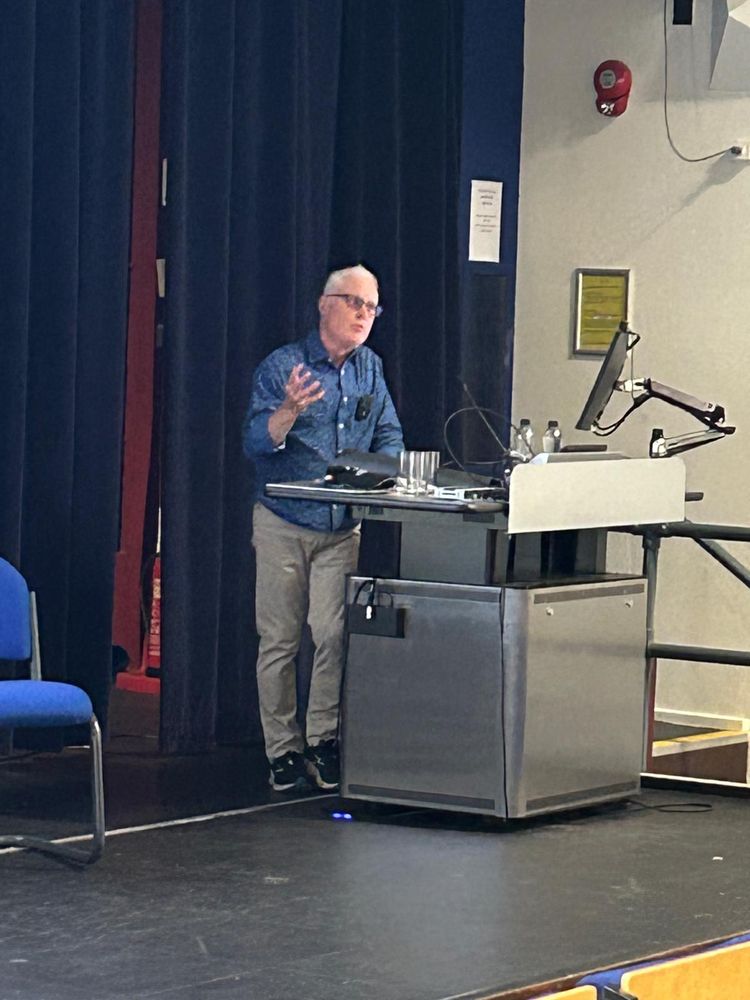 Professor John Goodby speaking from behind a metal podium.