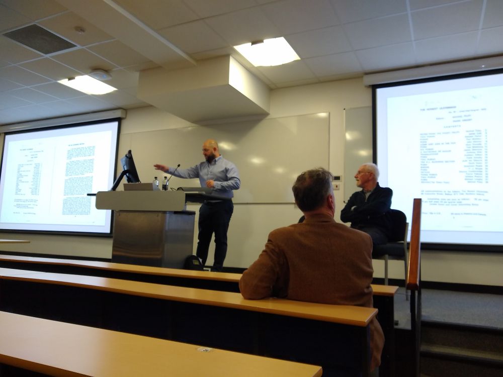 Dr James Gallacher, a white man dressed in dark trousers and medium blue shirt, speaking from the lectern in a modern lecture room. Micheal Foley is sitting to his left on the stage. Either side of them projections from Dr Gallacher's presentation are visible but overexposed.
