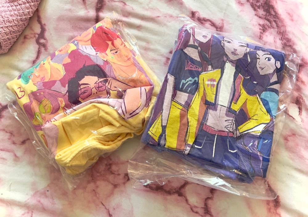 A photo of two plastic-wrapped Kpop Demon Hunters fan tshirts. The yellow one on the left is for the Saja Boys; the purple one on the right is for Huntrix. 