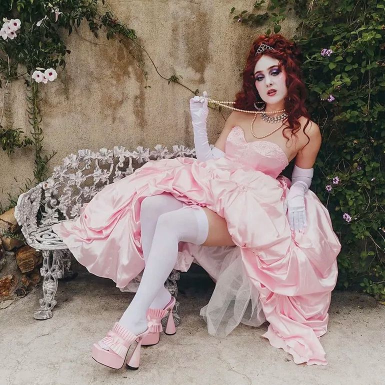 Chappell Roan sitting on a bench wearing a baby pink puffy dress, baby pink heels and white socks 