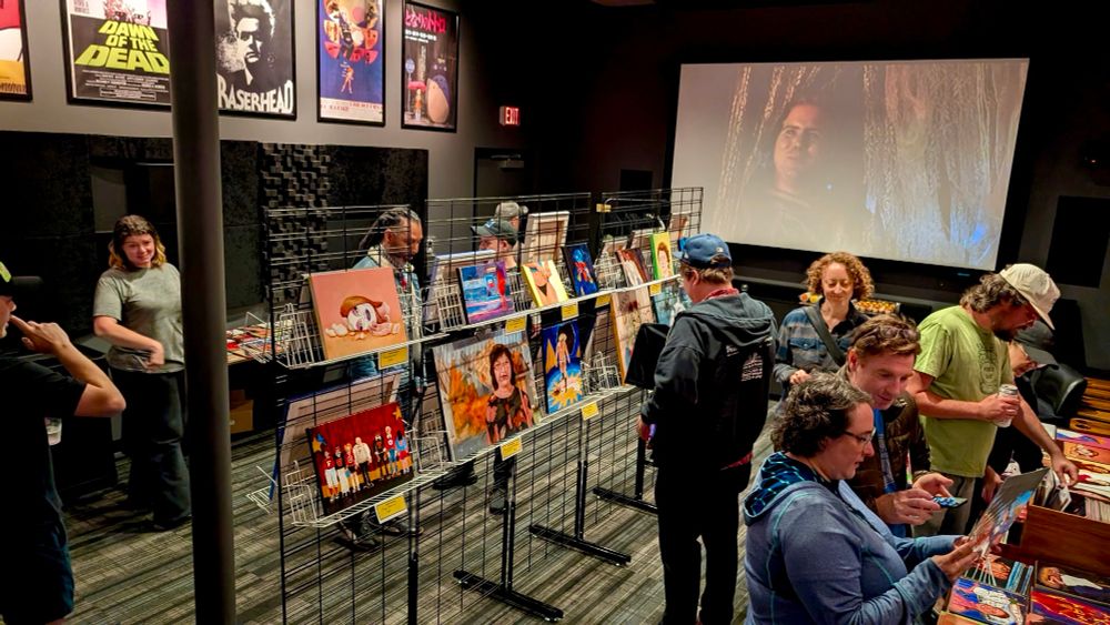 A photo of an art show in the miniplex at Movie Madness. Moments before the chaos hit. People viewing paintings in the center of the room with night of the demons playing on the big screen!