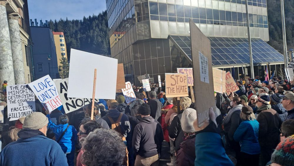 Protesters with signs in front of the Alaska State Capitol as sun shines and is reflected on court house.