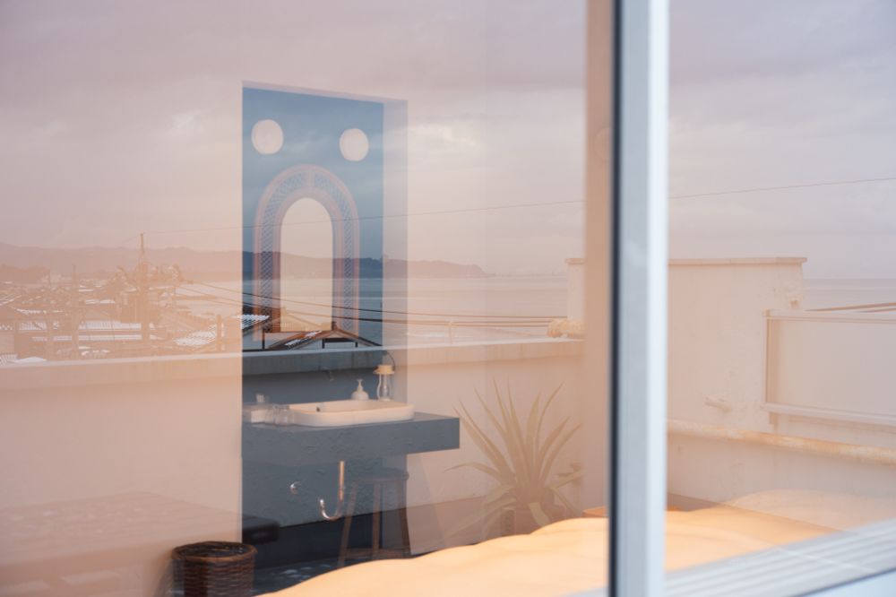 テラスから中の部屋を写した写真。部屋の中には洗面台が見えていて、反射で、海と家々の屋根がうつっている。