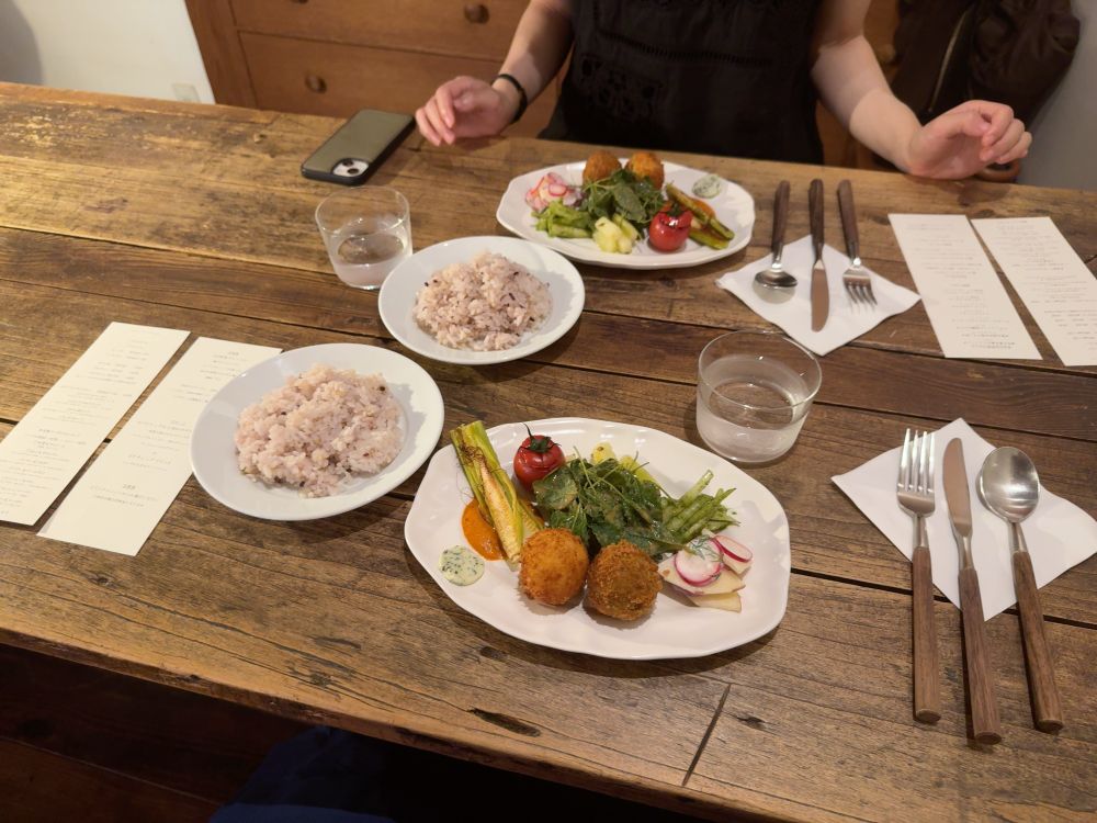 木のテーブルにコロッケや野菜が乗った平皿、ご飯が乗った平皿などがある。右にはスプーン、ナイフ、フォーク。左にはメニューの紙がある。
