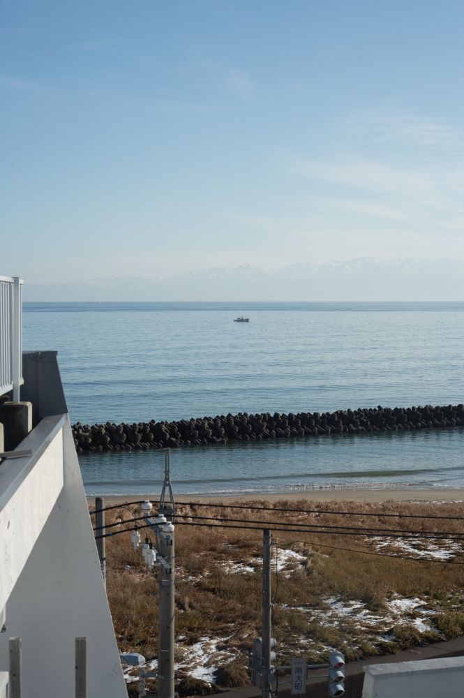 屋上に続く外階段から撮った晴れの富山湾の写真。左に屋上の柵、正面は海。一隻の船と、奥に立山連峰がうっすらと見える。