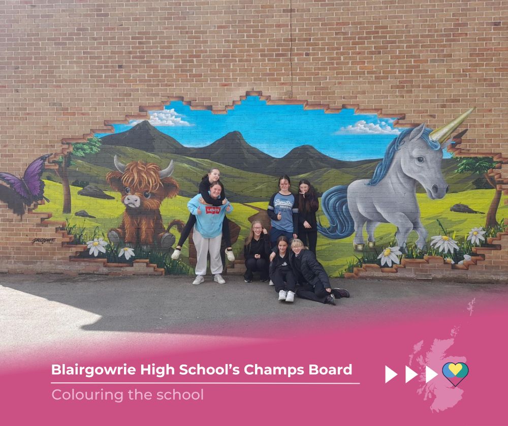 A group of young people gather in front of a colourful mural on a brick wall picturing rolling hills with a highland cow and a unicorn standing in the field. Flowers line the grass and a purple butterfly flies out of the image. 