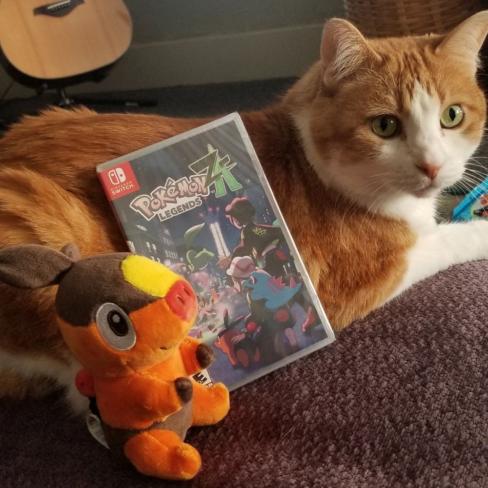 A photo of my cat Nikki, who is facing to the right posting for the camera while a copy of Pokemon Legends Z-A for the Switch is leaning up against her side. Nikki is an orange and white cat with green eyes. A plushie that came with my preorder is also sitting next to her, and it is of the pokemon Tepig. 