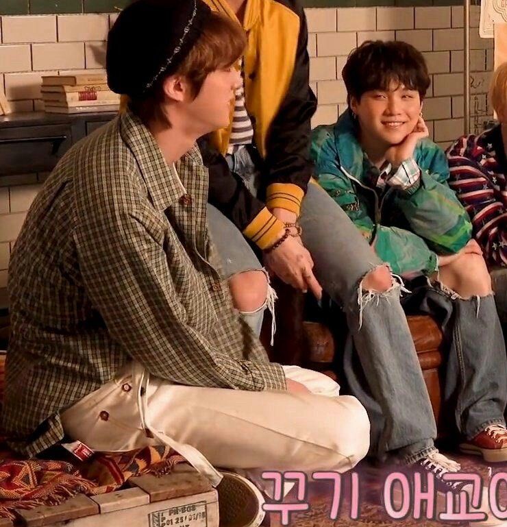 Yoongi smiling at Jin. BTS are all sitting down at a BTS photo shoot. Jin has brown hair and is wearing a beret, button down, light beige pants and sneakers. Yoongi has black hair, a green jacket, ripped jeans, and maroon sneakers.