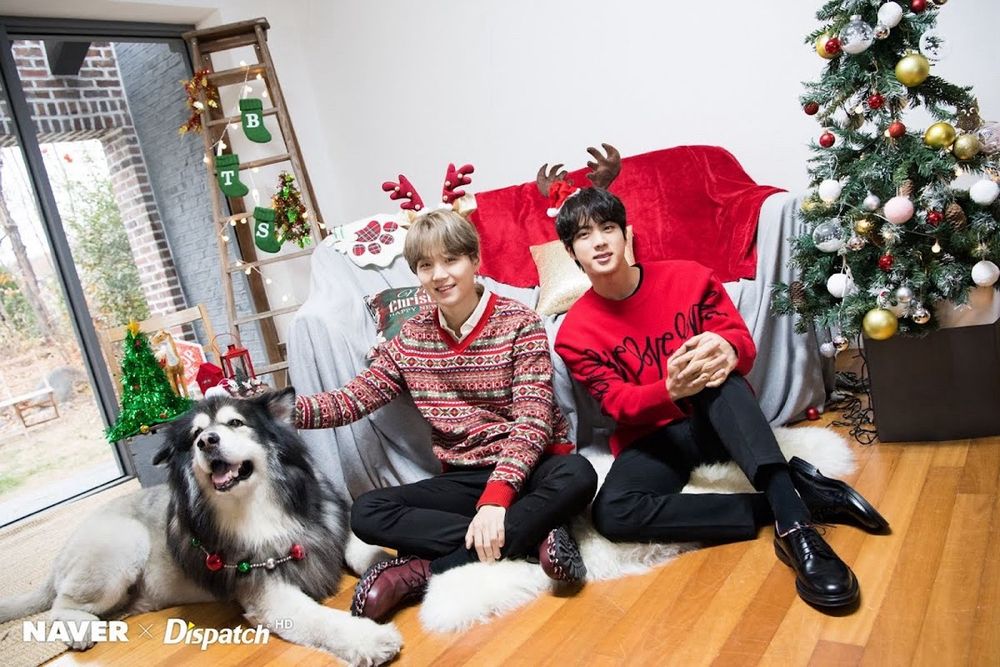 Yoongi and Jin are sitting in the floor in a room decorated for Christmas. Yoongi is letting a big dog. They are wearing reindeer antler headbands and Christmas sweaters with black slacks and dress shoes. Yoongi has honey blond hair and Jin has black hair.