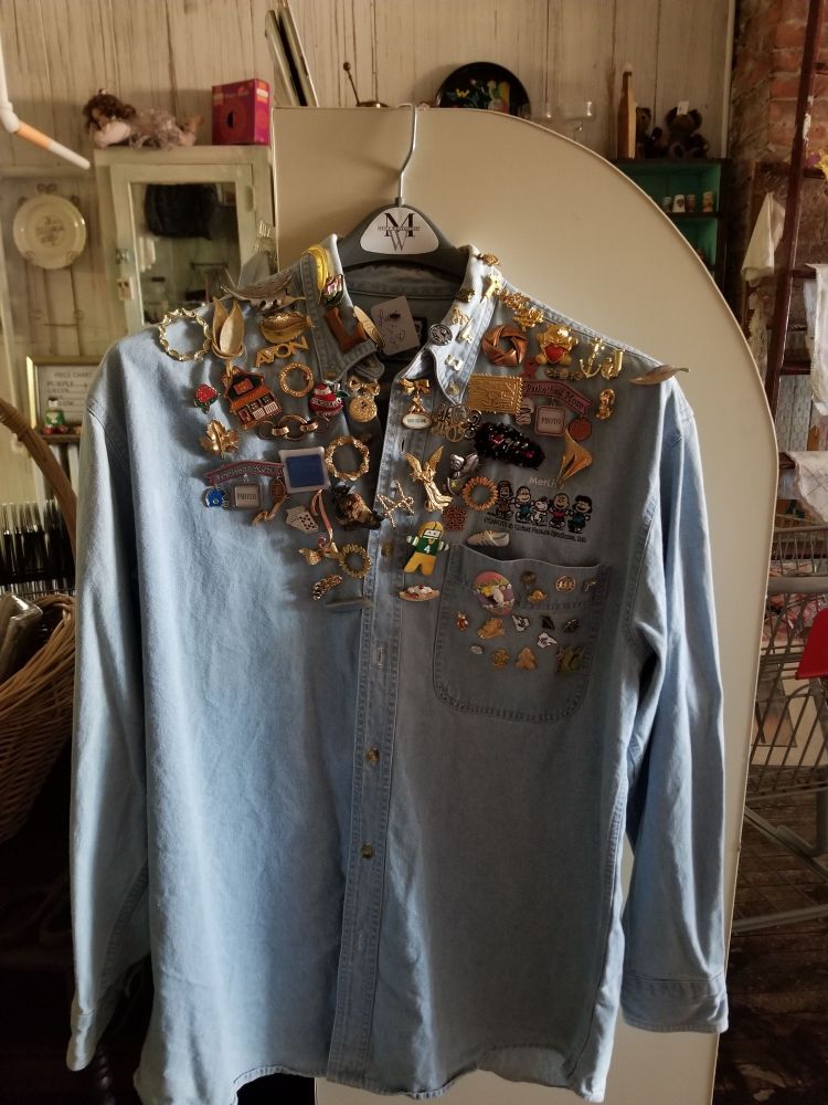 A long sleeved denim button-up shirt with a left breast pocket. The collar and front of the shirt, including the pocket, is covered in brooches and pins, mostly old avon and costume jewelry. 