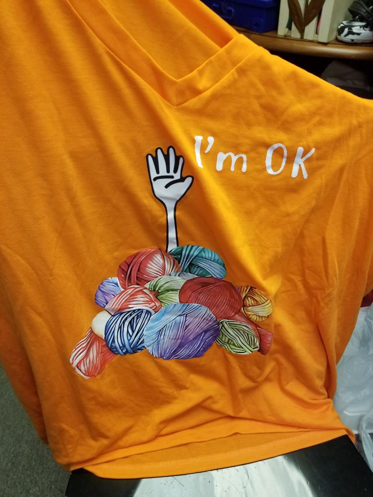 A bright orange women's shirt. A large image on the front of the shirt features a pile of yarn with a cartoon hand sticking out of it as if waving. The caption, in white letters to the right of the hand, reads: "I'm OK"
