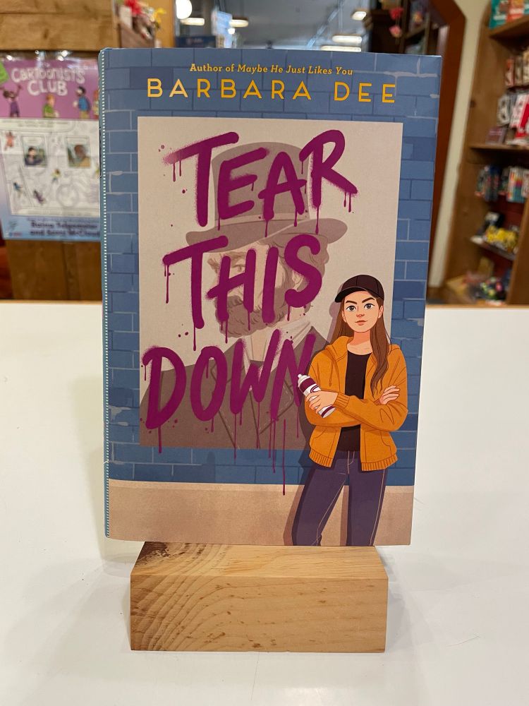 Cover of Tear This Down by Barbara Dee shows a tween girl wearing a black beanie and holding a can of spray paint. The book title looks like fresh paint and is over the profile of a man wearing a hat. 