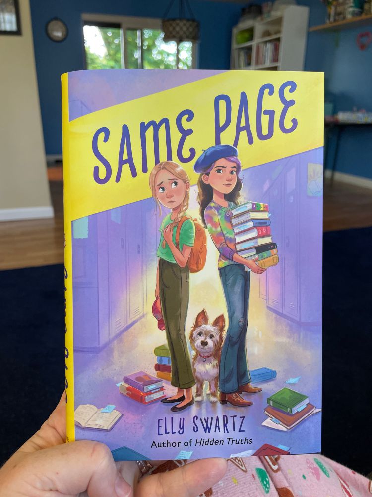 Same Page by Elly Swartz. Two 6th grade girls standing back to back in a school hallway with a small dog between their legs facing the viewer. One girl is holding a large stack of books. 