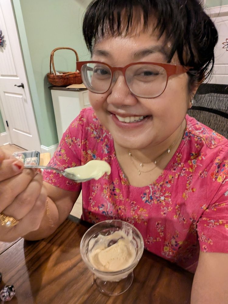 An Asian woman in red glasses and a pink dress smiles while holding up a spoonful of ice cream.