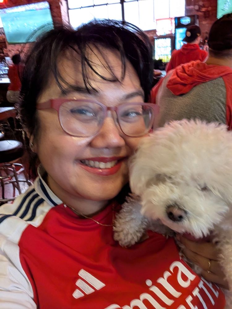 An Asian woman in red glasses and the 24/25 Arsenal home kit snuggles a fluffy white dog and smiles for the camera.