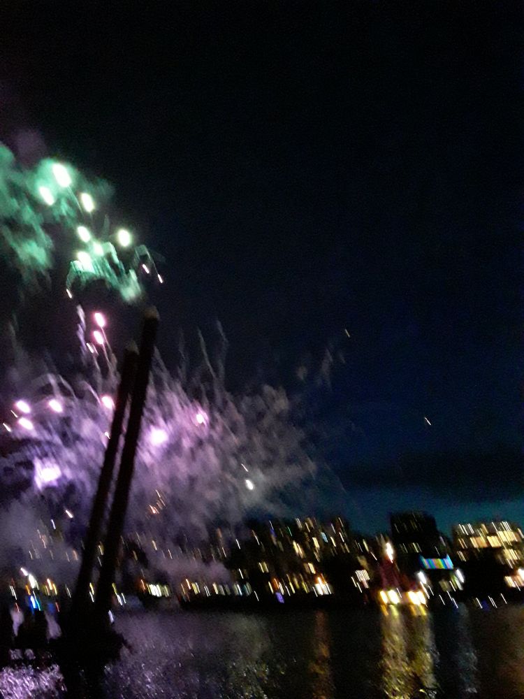 Purple and green fireworks behind a boat moring.city lights from across the river reflected in the water.