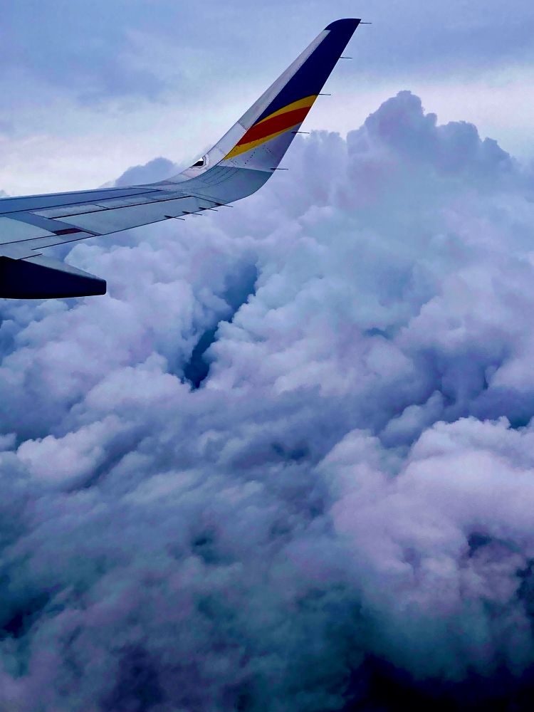 Blue grey cloudscape from the window of a plane with a bit of the silver wing showing a dash of red and blue at its tip