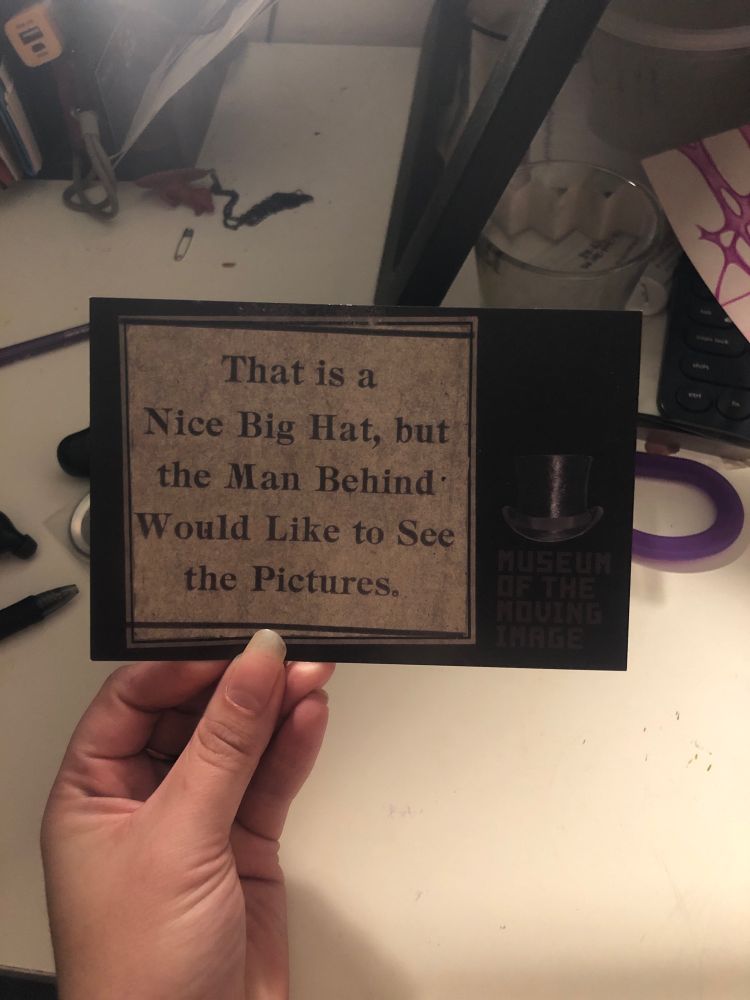 photo of a hand holding a post card that has art of a top hat and the text "That is a Nice Big Hat, but the Man Behind Would Like to See the Pictures"