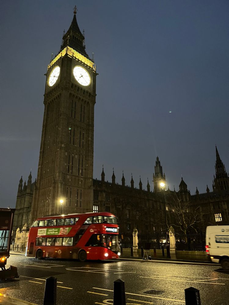 A view of the Houses of Parliament with a red London bus in the foreground. The sky is dark, it’s early morning.